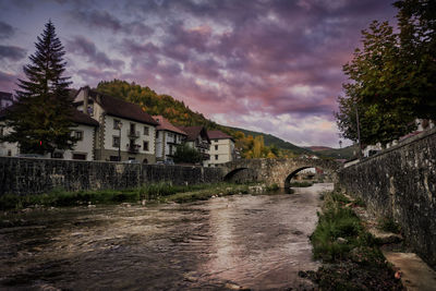 Houses by river and buildings against sky at dusk