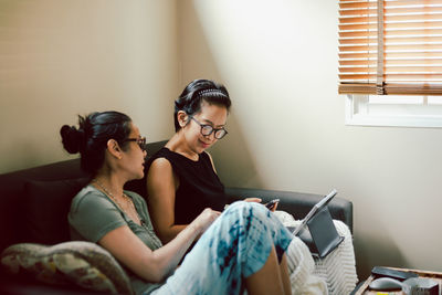 Side view of two woman sitting on sofa at home using laptop.