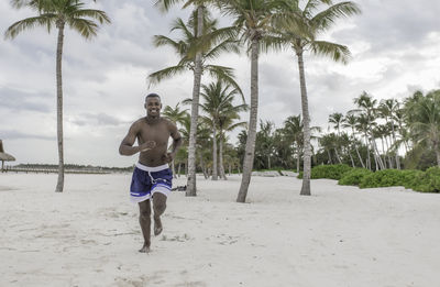 Smiling young black guy shirtless running along the beach with palm trees and resort