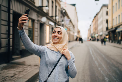 Smiling young woman taking selfie from smart phone while standing on street in city