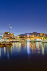 Illuminated buildings by river against blue sky at night