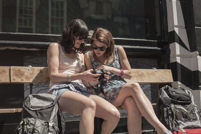 Curious woman looking at camera held by female friend while sitting on bench in city