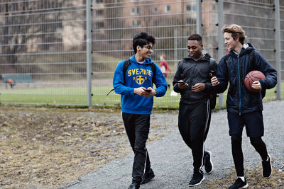 Happy friends using mobile phones while walking on street against sports court