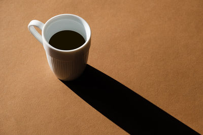 A cup of coffee on a brown background casts a long shadow.