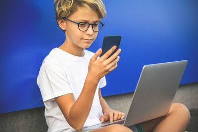 Boy sitting with laptop while using smart phone against blue background