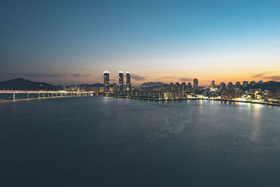 Illuminated city by sea against sky at dusk