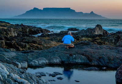 Rear view of young man kneeling on rock by sea against sky during sunset