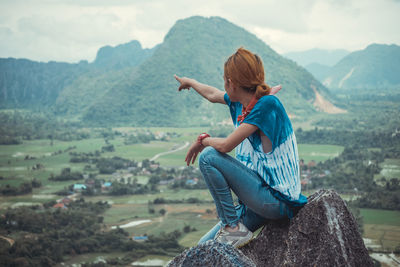 Side view of mid adult woman pointing at mountains while sitting on cliff
