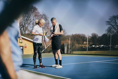 Senior man and woman with rackets talking while standing at tennis court