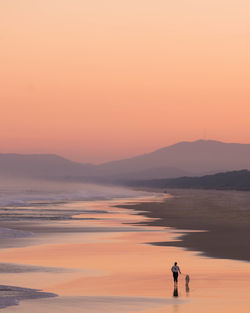 Woman standing on sea shore against orange sky