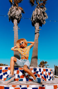 Full length of woman sitting by tree against blue sky