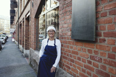 Portrait of smiling baker leaning on brick wall outside bakery