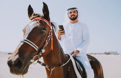 Man using mobile phone while horseback riding against sky