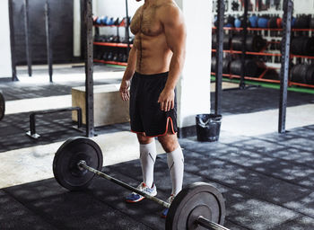 Low section of shirtless man standing by barbell in gym