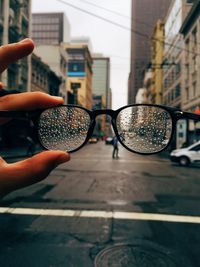 Cropped image of hand holding wet eyeglasses against city street