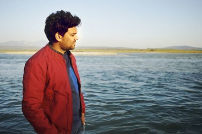 Young man looking away while standing by sea against sky