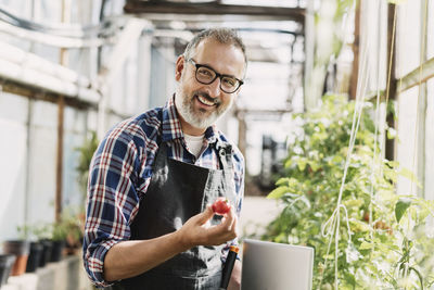 Smiling gardener with digital tablet holding tomato in greenhouse