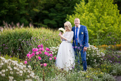 Groom making face while bride holding bouquet while standing amidst plants at park