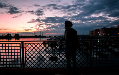Silhouette man standing by railing against river in city during sunset