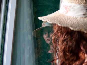 Woman wearing hat while looking at her reflection in window frame
