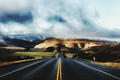 Empty road leading towards mountains against cloudy sky