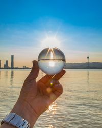 Hand holding crystal ball against sky during sunset