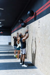 Full body side view group of hispanic sportspeople in activewear standing near wall with heavy medical balls during weightlifting workout in gym