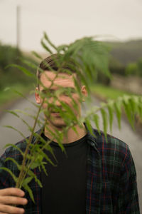 Portrait of young man holding plant while standing on road against sky