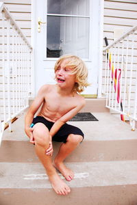 Full length of shirtless boy crying while sitting on steps against door