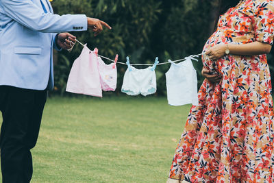 Midsection of pregnant woman with man holding baby clothing on field