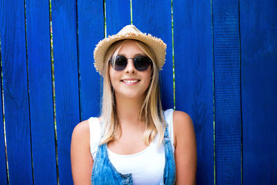 Portrait of smiling teenage girl wearing sunglasses and hat against fence