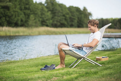 Full length side view of mature man using laptop on deck chair at lakeshore