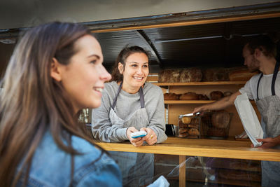 Smiling saleswoman accepting payment from customer while colleague packing bread at concession stand