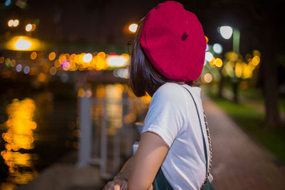 Side view of young woman standing at railing against canal in illuminated city during night