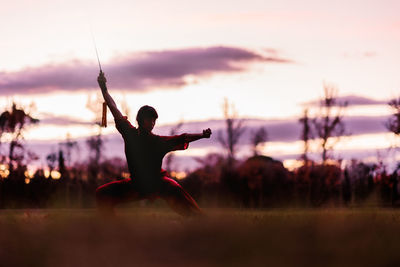 Side view of woman with arms outstretched on field against sky during sunset