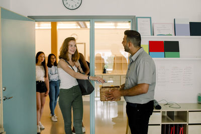 Smiling teenage girl giving phone to teacher while walking in classroom