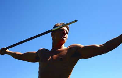 Low angle view of young shirtless man throwing javelin against clear sky