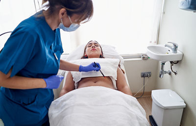 From above beautician in uniform and face mask drawing with pencil preparing to do laser lipolysis to female client during work in aesthetic clinic