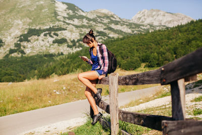 Female hiker using phone while sitting on fence against mountains