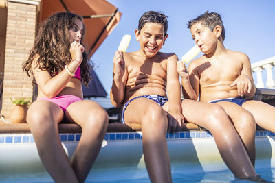 Friends eating popsicle while sitting on poolside against clear sky