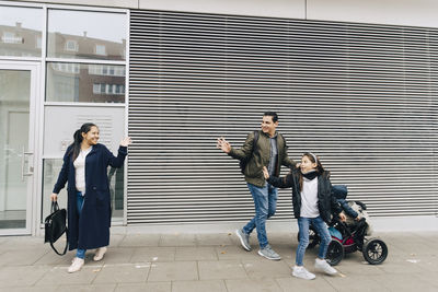 Father and daughter waving to mother while walking on sidewalk with stroller in city