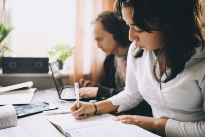 Teenage girl writing in book while friend using laptop at home