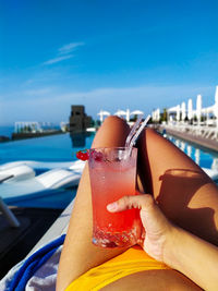 Close-up of hand holding drink at swimming pool against sky