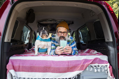 Concentrated mature male browsing smartphone while lying in mini camper with french bulldog during road trip in countryside