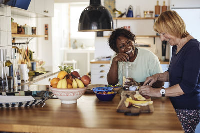 Happy multiracial female friends making fruit smoothie together on kitchen counter at home