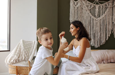 Boy holding milk bottle while mother sitting on bed