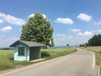 Road by trees and houses on field against sky