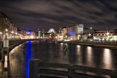 Illuminated bridge over river by buildings against sky at night