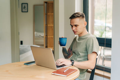 Young man drinking coffee reading social media from laptop screen while sitting at table at home.