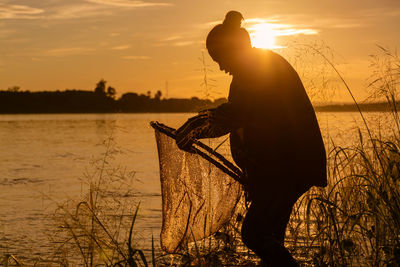 Silhouette of woman fishing while standing by lake during sunset